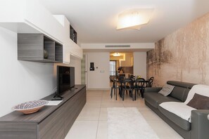 A modern living room with a flat-screen TV, a sofa, a dining area with chairs, and a kitchen in the background.