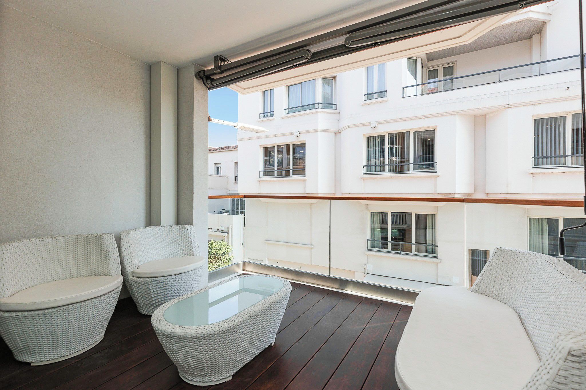 A balcony with wicker furniture, a glass table, and a view of a white building with balconies.
