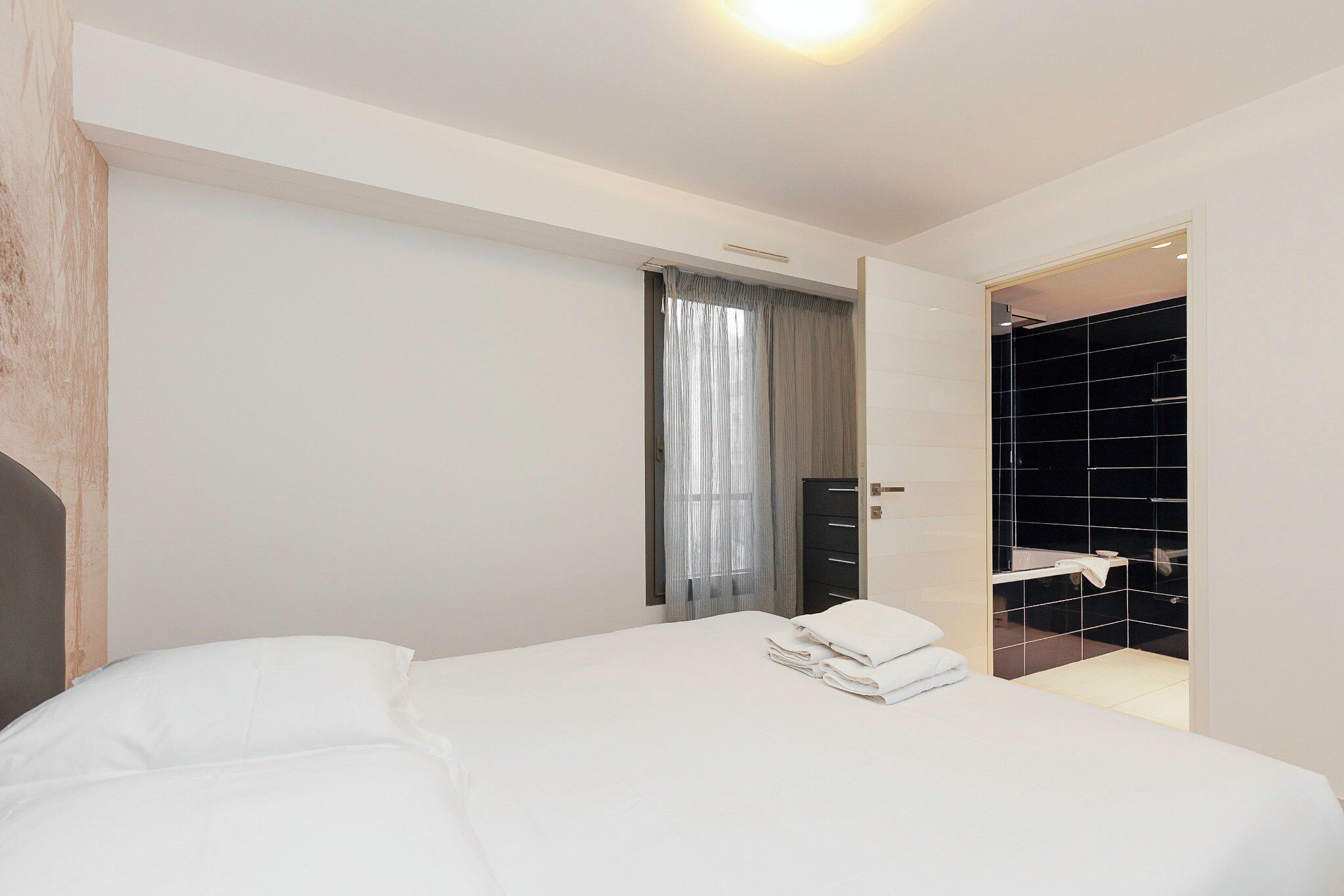 A modern hotel room with a neatly made bed, a bathroom with a black tiled shower, and a large window with sheer curtains.