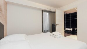 Een moderne hotelkamer met een net opgemaakt bed, een badkamer met een douche met zwarte tegels, en een groot raam met dunne gordijnen.
