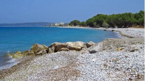 A pebble beach with clear blue water, rocky shoreline, and a forested area in the background. - Smart Accomodation Rhodes by GHH (Rhodes)