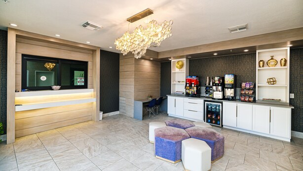 A modern reception area with a wooden reception desk, a seating area with purple and white cushions, and a counter with various amenities. - Palace Inn IAH Houston Bush Intercontinental Airport (Humble)