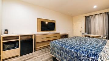 Habitación de hotel con cama, televisor montado en la pared, escritorio con silla y una pequeña zona de kitchenette.