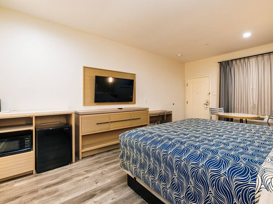 Habitación de hotel con cama, televisor montado en la pared, escritorio con silla y una pequeña zona de kitchenette.