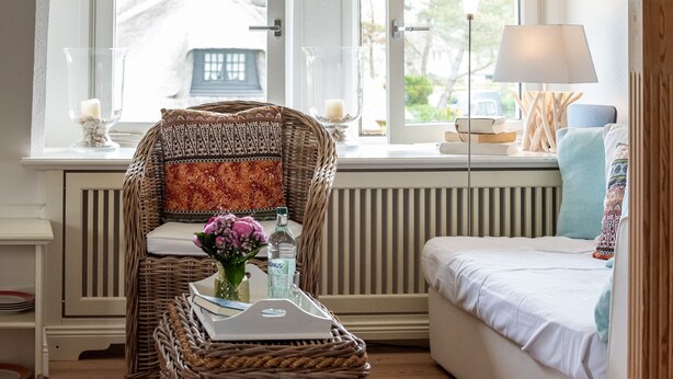 A cozy room with a wicker chair, a bedside table with a vase of flowers, a lamp, and a view of a house through the window. - Cozy 3-room apartment on approx. 55 m² for 4 persons. (Wenningstedt-Braderup (Sylt))