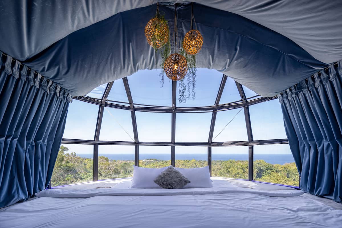 A bed with white linens and a single gray pillow, set against a backdrop of a clear blue sky and a view of the ocean.