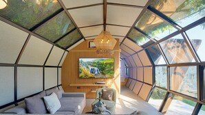 A modern, spacious room with a large TV, comfortable seating, and large windows offering a view of the outdoors. - Airship Bali by Alex Villas (Pecatu)