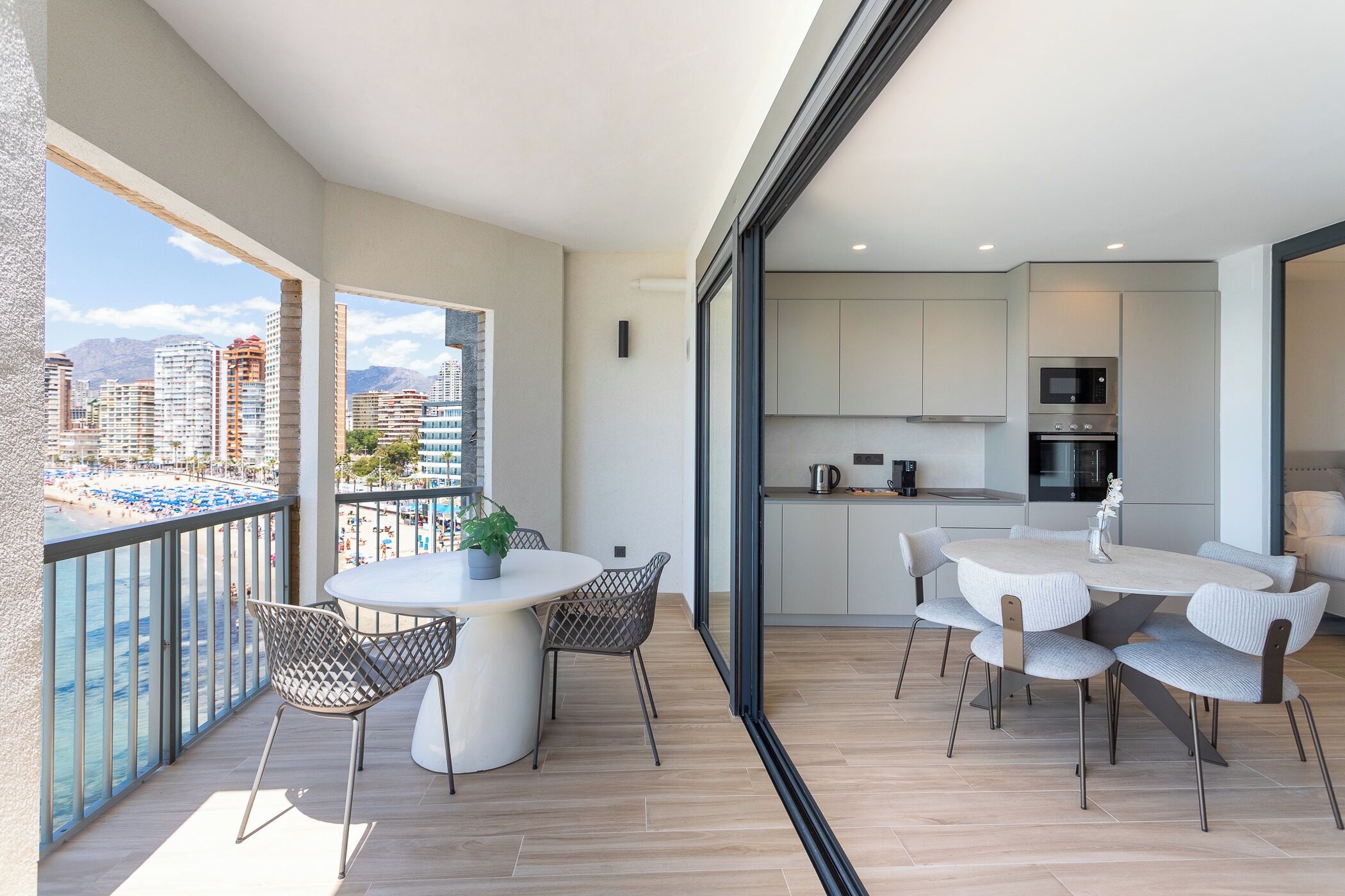 Een modern appartement met balkon en uitzicht op zee, een keuken met moderne apparatuur, en een eethoek met een ronde tafel en stoelen.