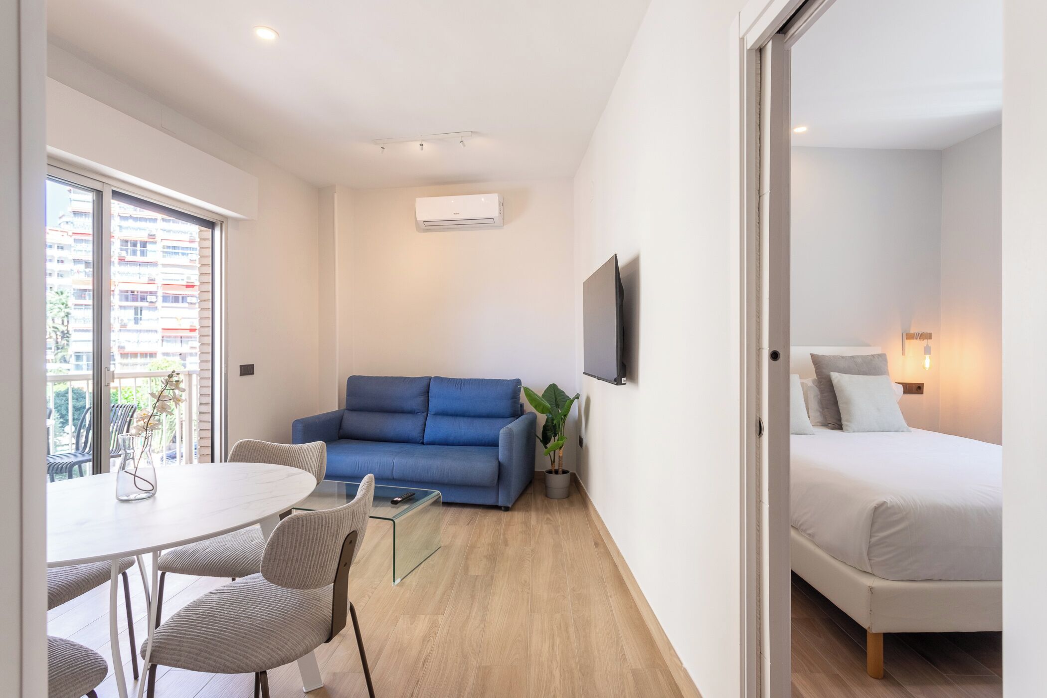 Quarto moderno com sofá azul, cama branca, mesa de jantar com cadeiras e televisão na parede.
