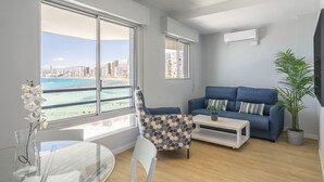 A modern living room with a large window offering a city view, a blue sofa, a white coffee table, and a potted plant. - Arenal Suites Benidorm (Benidorm)