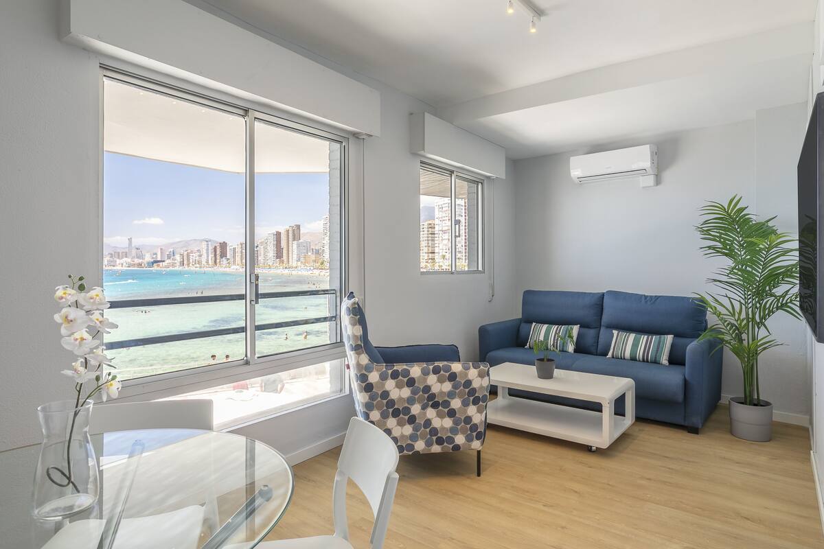 Uma sala de estar moderna com uma janela grande que oferece vista para a cidade, um sofá azul, uma mesa de centro branca e uma planta em vaso.