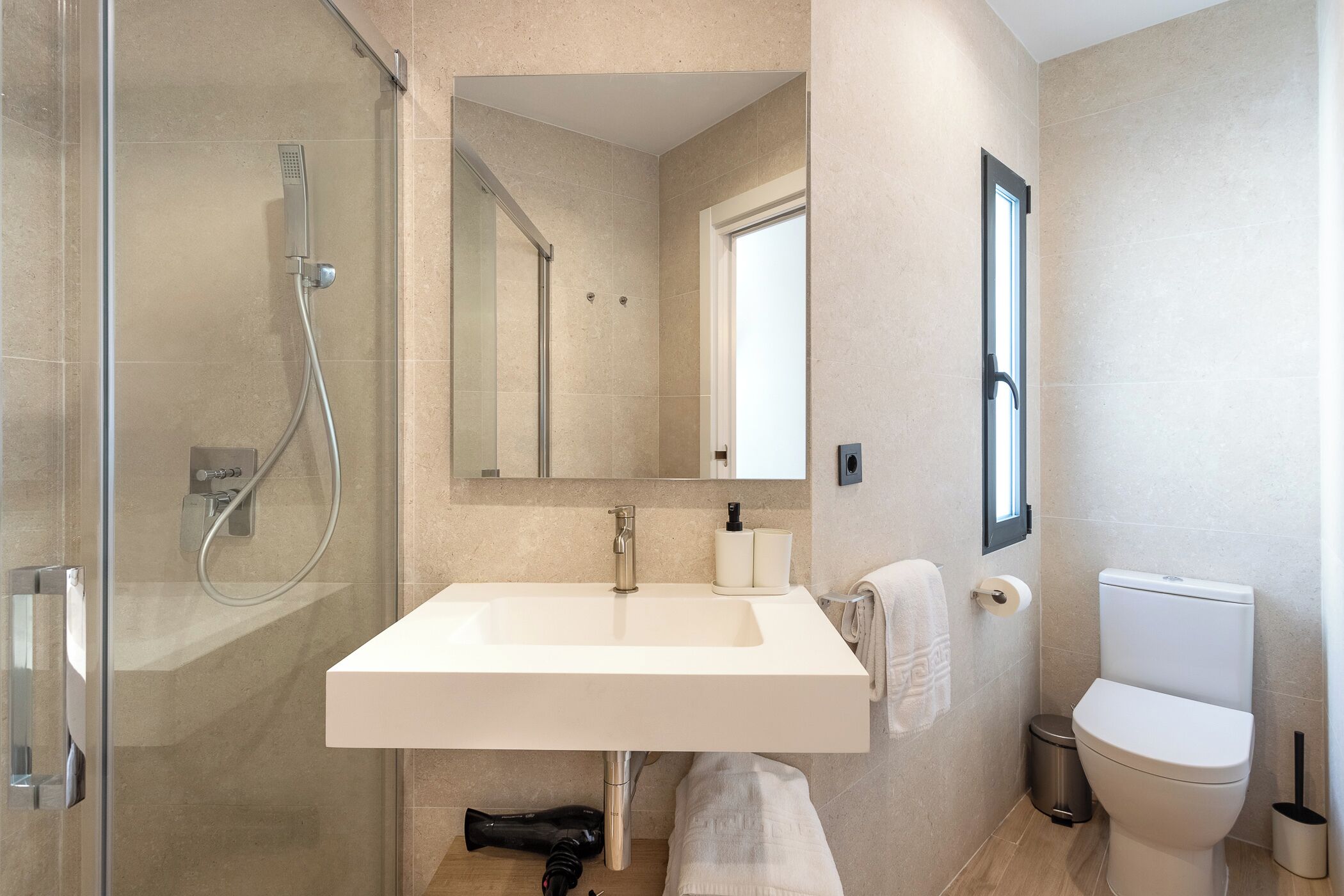 Un baño moderno con ducha envuelta en vidrio, lavamanos blanco, inodoro y espejo sobre el lavamanos.