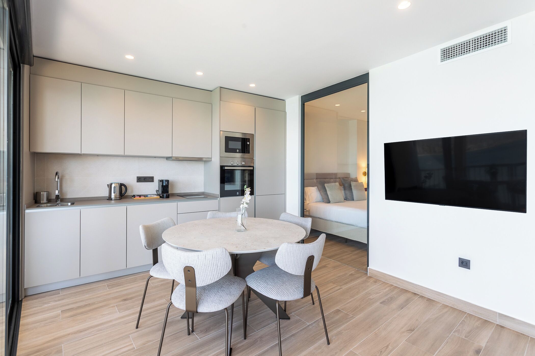 A modern kitchen with white cabinets, a round dining table with four chairs, a built-in oven, and a flat-screen TV mounted on the wall.