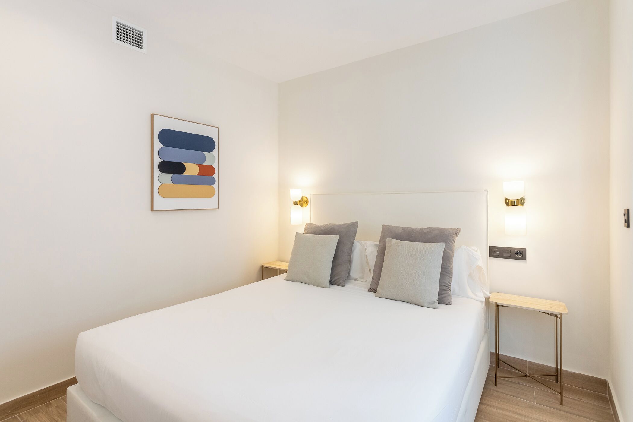 Una cama bien hecha con edredón blanco, dos mesitas de noche con lámparas y una obra de arte en la pared.