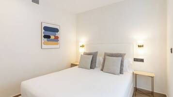 Una cama bien hecha con edredón blanco, dos mesitas de noche con lámparas y una obra de arte en la pared.