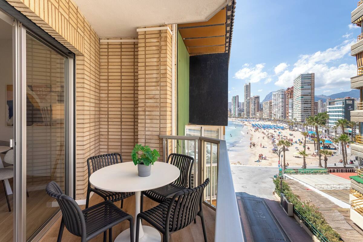 Uma varanda com mesa e cadeiras, uma planta em vaso e vista para a praia e o horizonte urbano.
