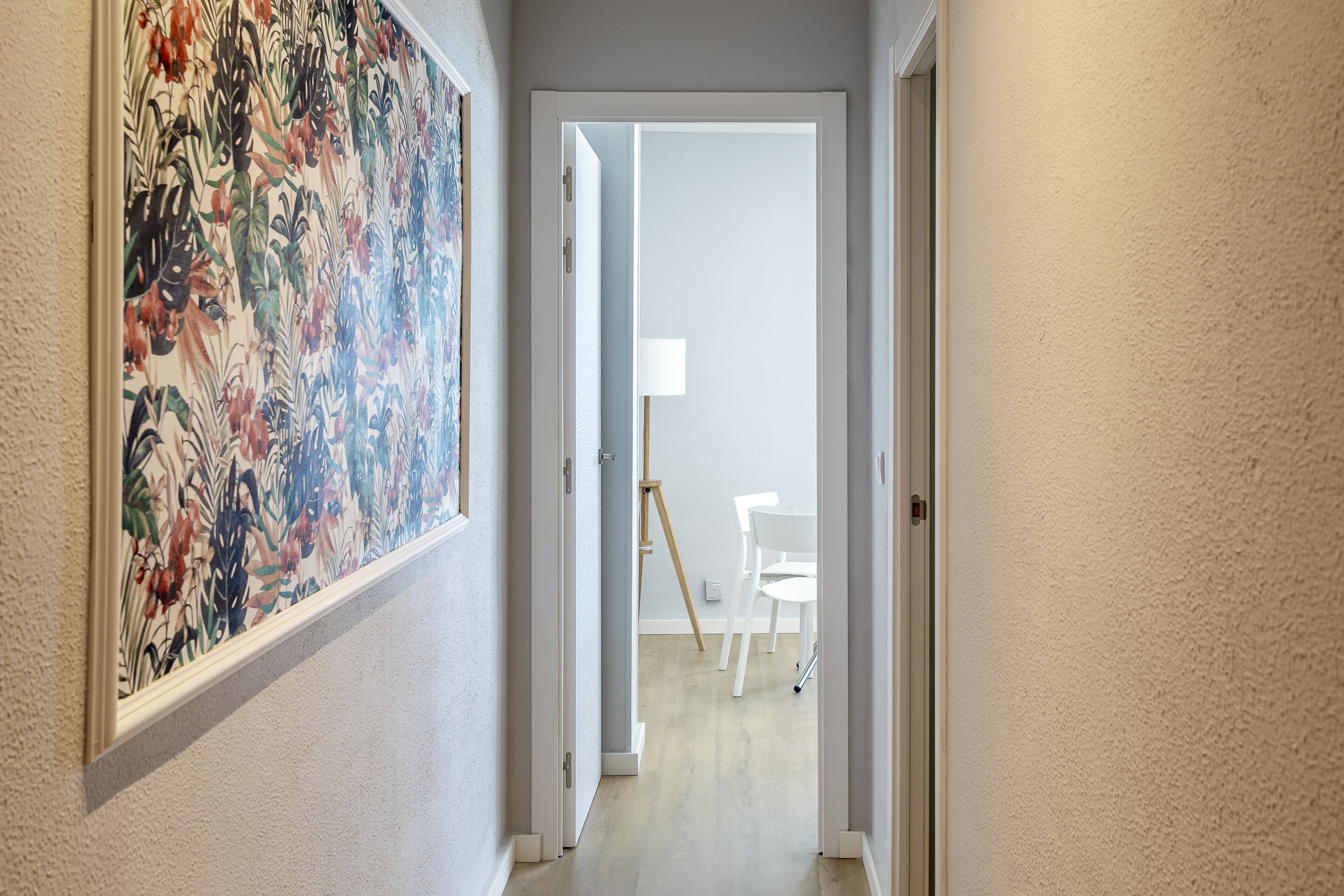 Un pasillo con una obra de arte floral enmarcada a la izquierda, una puerta blanca al final, y una silla y una lámpara blancas al fondo.