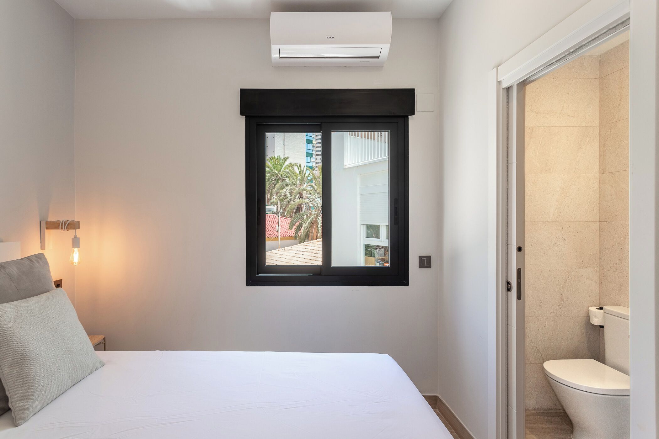 A bedroom with a bed, a window, an air conditioner, and a view of the outside.