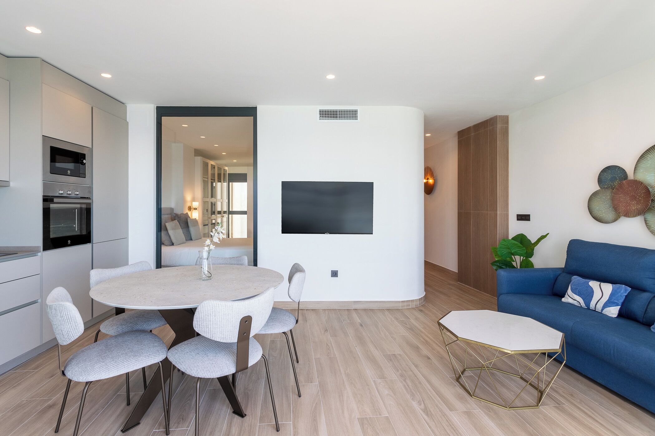 A modern kitchen with built-in appliances, a dining area with a round table and chairs, a flat-screen TV mounted on the wall, and a blue sofa in the living area.