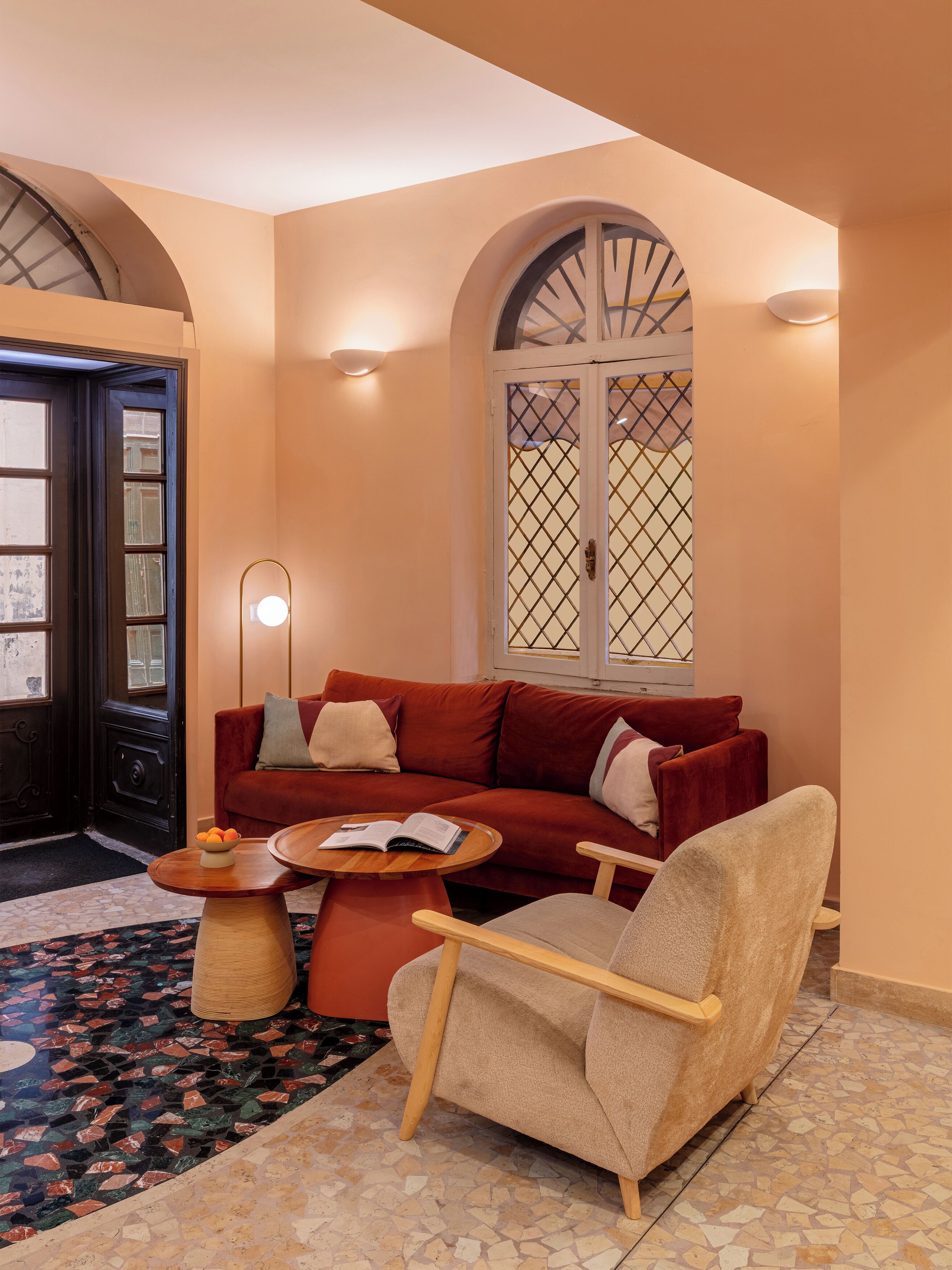 Una sala de estar acogedora con un sofá rojo, una mesa de centro de madera y un sillón beige. Hay una alfombra estampada en el suelo y una lámpara de pared decorativa.