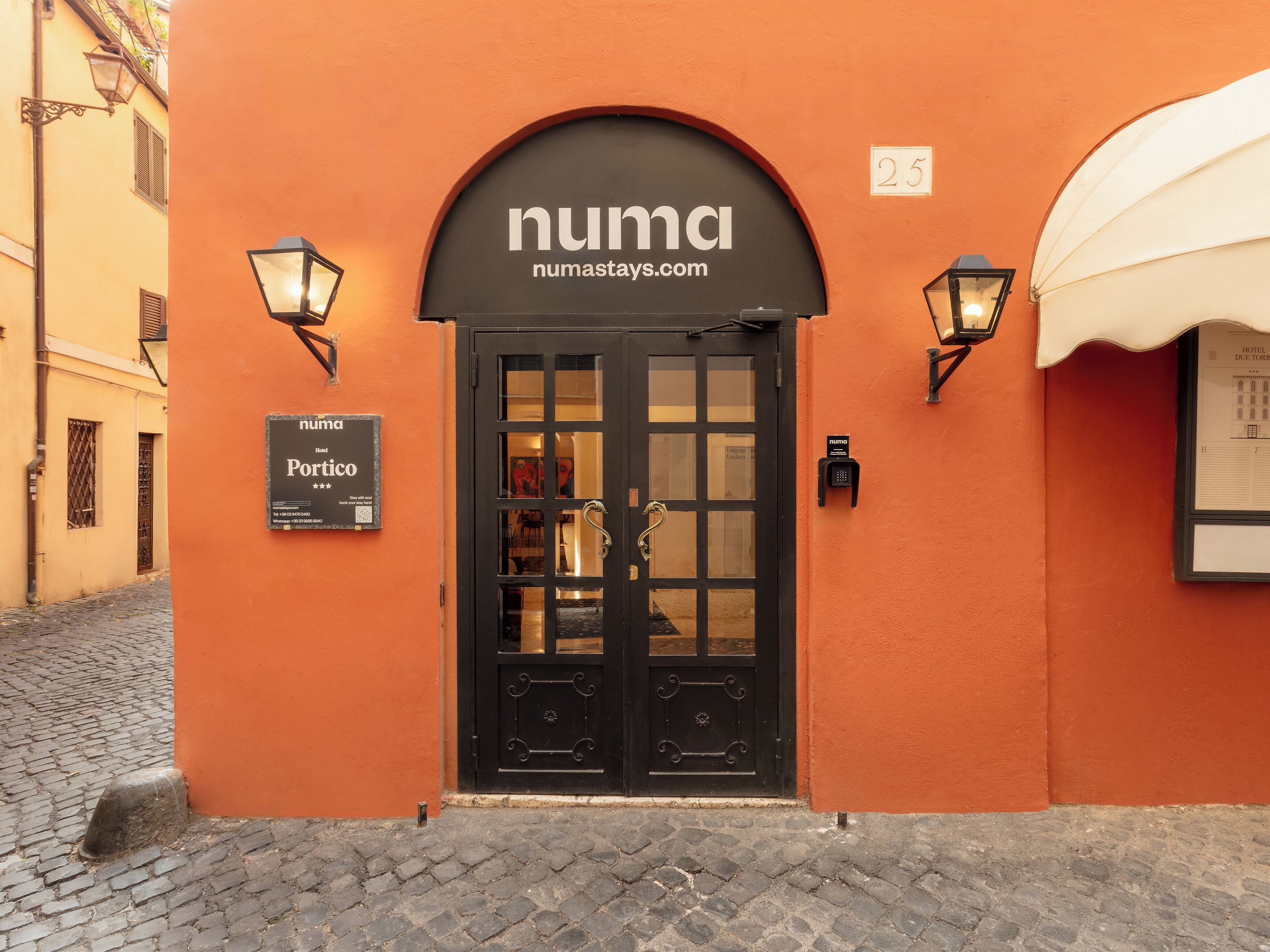 Entrada de hotel con una puerta negra, dos lámparas fijadas en la pared y un cartel sobre la puerta.