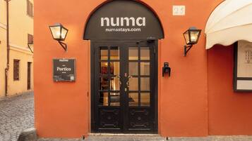 Entrada de hotel con una puerta negra, dos lámparas fijadas en la pared y un cartel sobre la puerta.