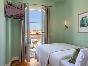 A hotel room with a bed, a small balcony with a table and chairs, a welcome sign, and a cityscape view.