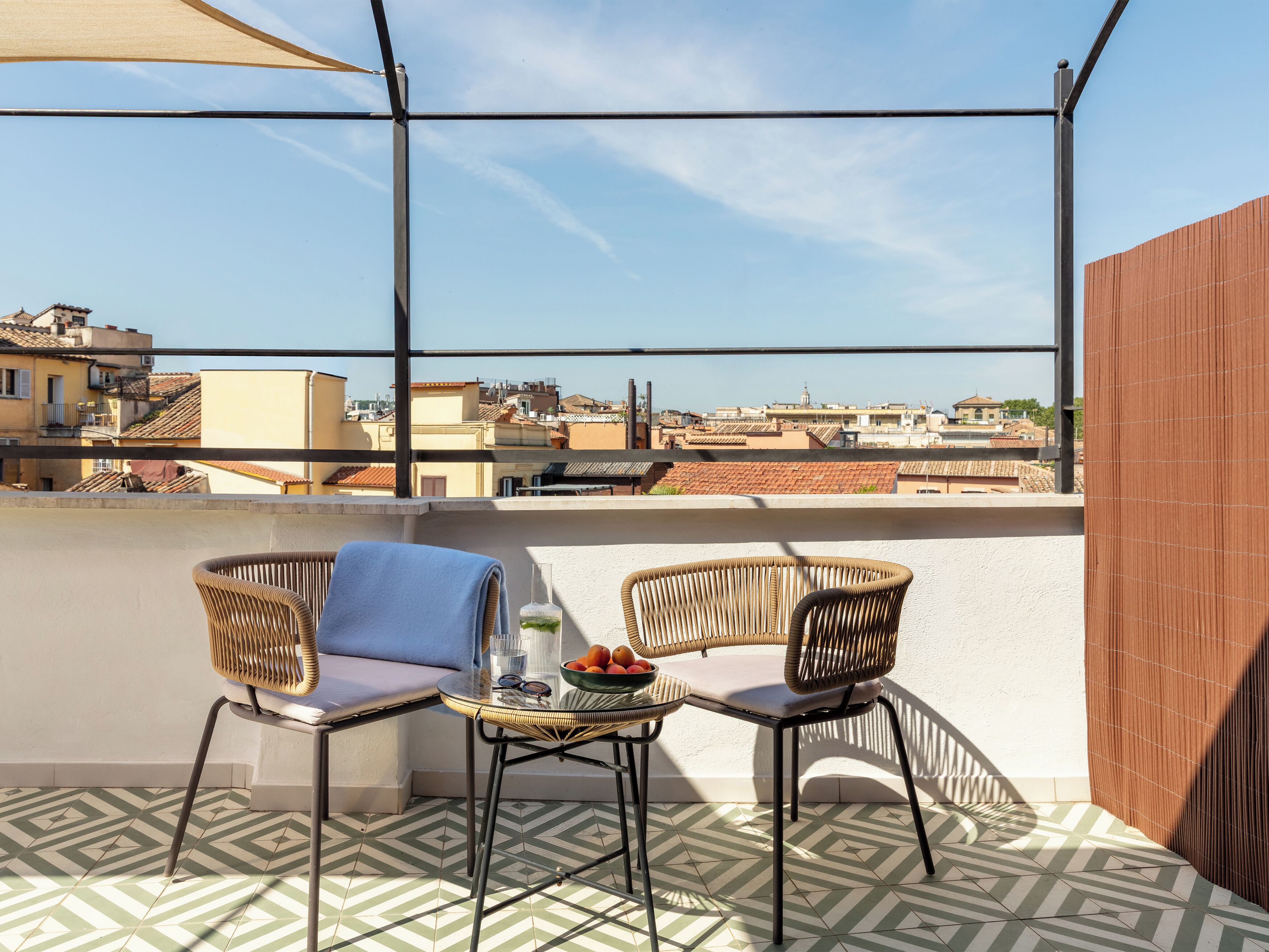 Una terraza en la azotea con dos sillas de mimbre, una mesita y un cuenco de fruta. La terraza tiene baldosas con diseños y vistas de la ciudad.