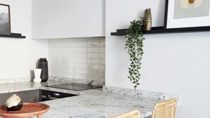 Una cucina con piano di lavoro in marmo, due sgabelli da bar in vimini, lampade a sospensione e una pianta in vaso appesa al muro.