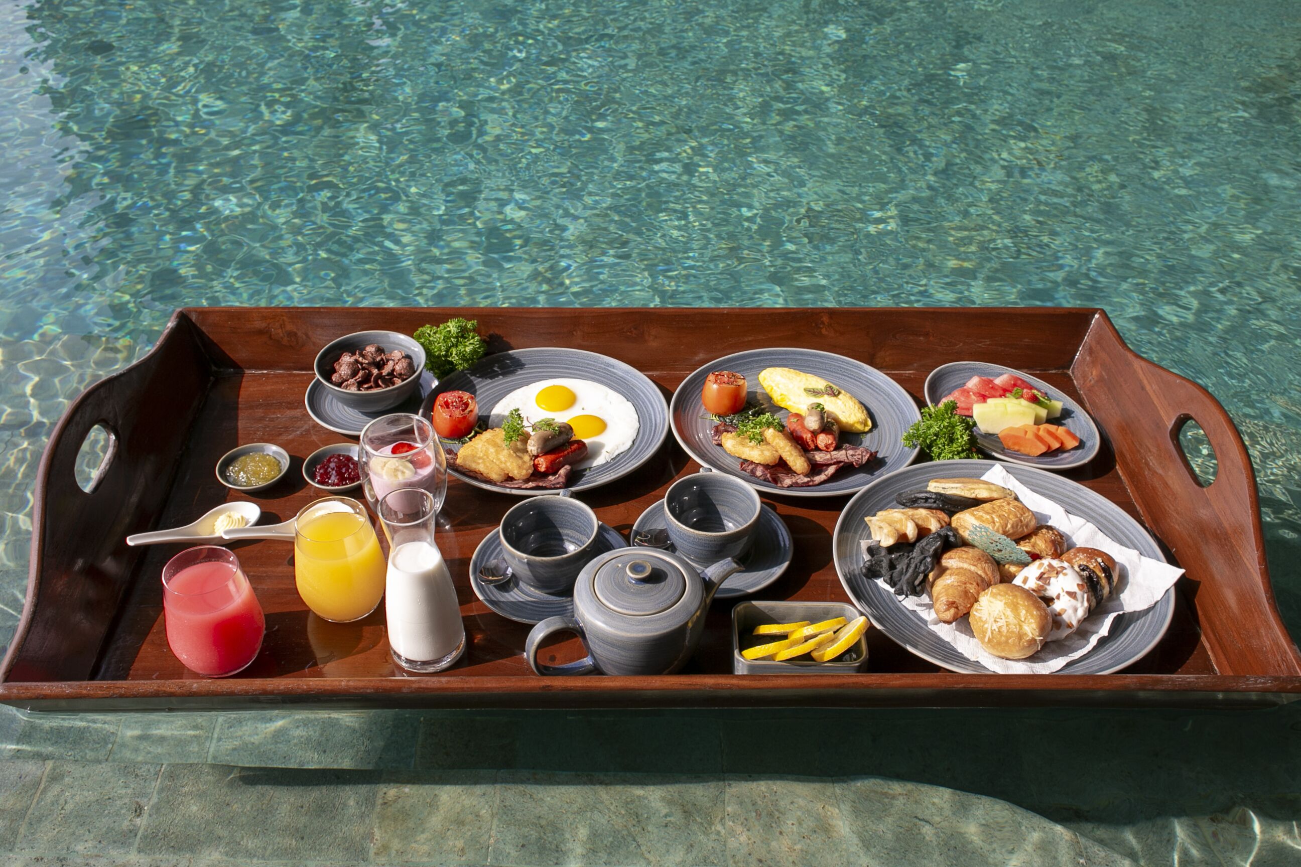 Un plateau en bois contenant divers aliments pour le petit-déjeuner, notamment des œufs, des toasts, des fruits et des boissons.