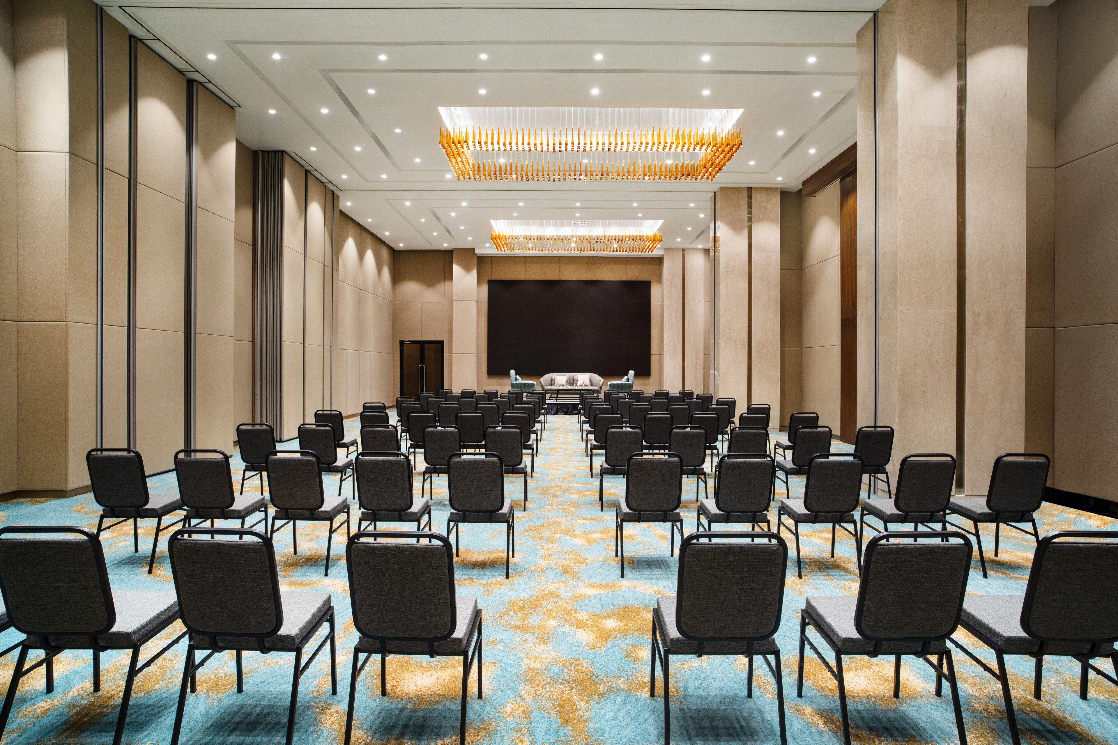 A conference room with rows of chairs facing a stage, beige walls, and a patterned carpet.