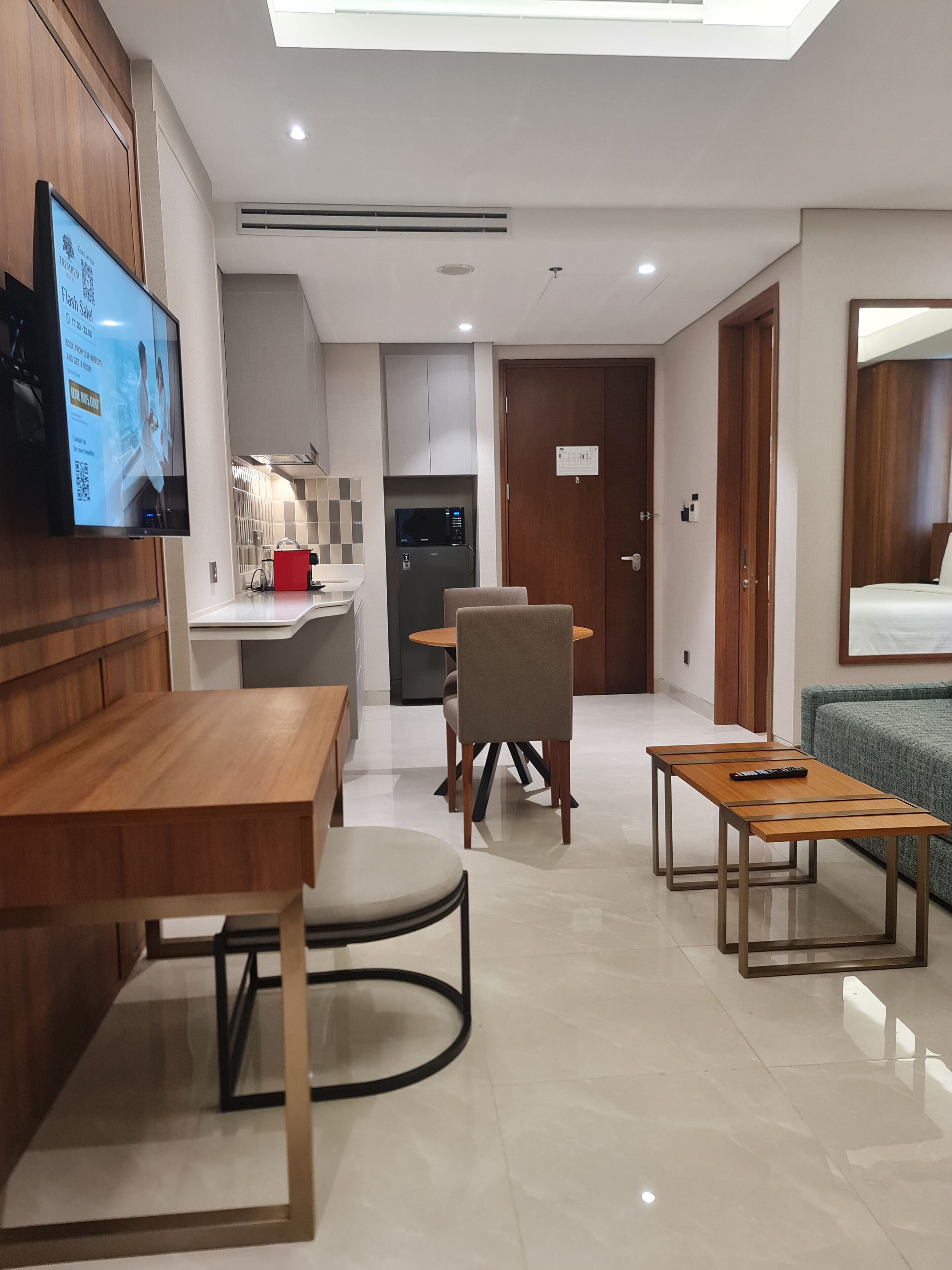 Een moderne hotelkamer met een houten eettafel, een bank, een aan de muur bevestigde televisie, een koelkast, en een deur met slot.