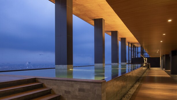 A modern indoor pool area with a view of the city skyline, featuring a long corridor with steps leading to the pool, and large pillars supporting the ceiling. - Trembesi Hotel (South Tangerang)