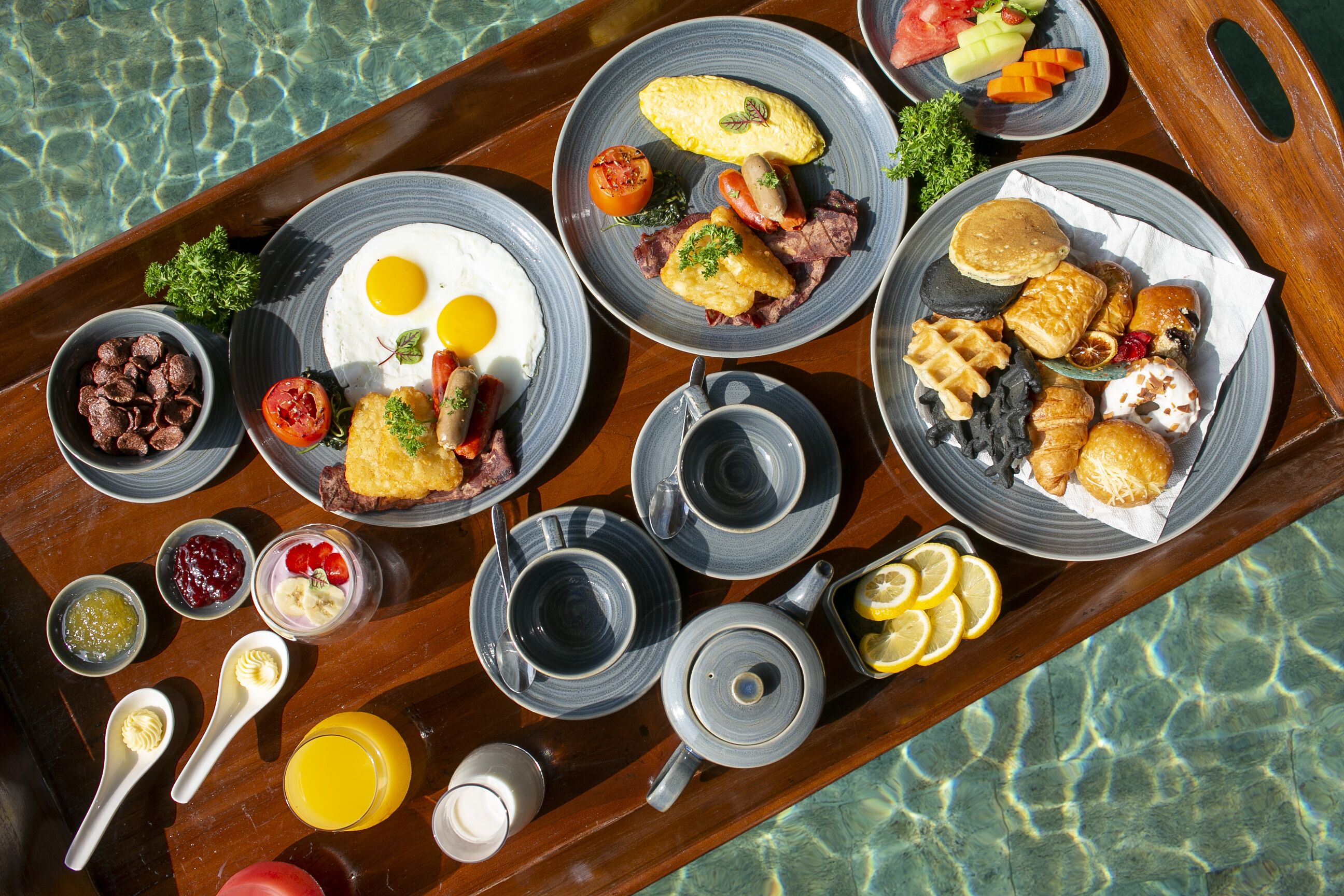 A breakfast tray with various dishes and drinks, including eggs, sausages, and toast, placed on a wooden table by a pool.
