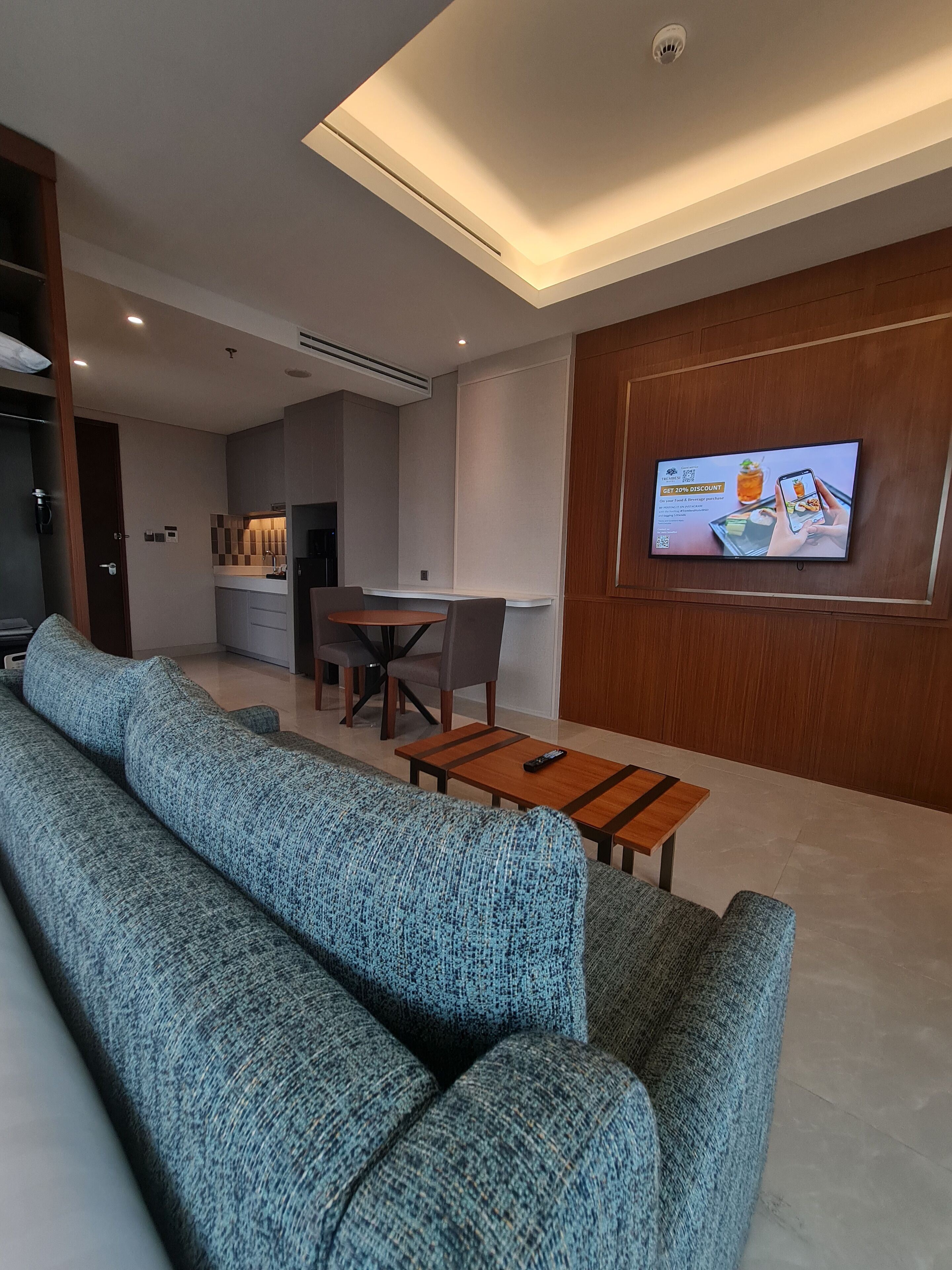 A modern living room with a sectional sofa, a dining area with a table and chairs, and a television mounted on the wall displaying an advertisement.