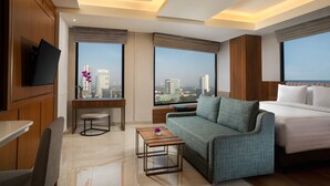 A modern hotel room with a large bed, a sofa, a small table, a TV, and a city view. - Trembesi Hotel (South Tangerang)
