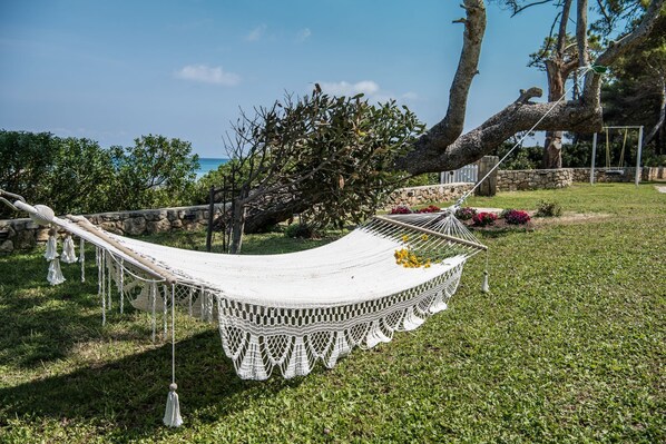 A hammock in a grassy outdoor area with a stone wall and trees in the background.