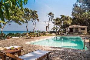 A pool area with a clear view of the sea, a small building, and a table with fruits and drinks.