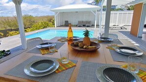Un espacio de comedor al aire libre con piscina, mobiliario blanco y plantas tropicales.