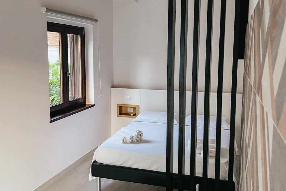 A bedroom with a single bed, a window with curtains, a bedside table, and a wall-mounted light fixture.
