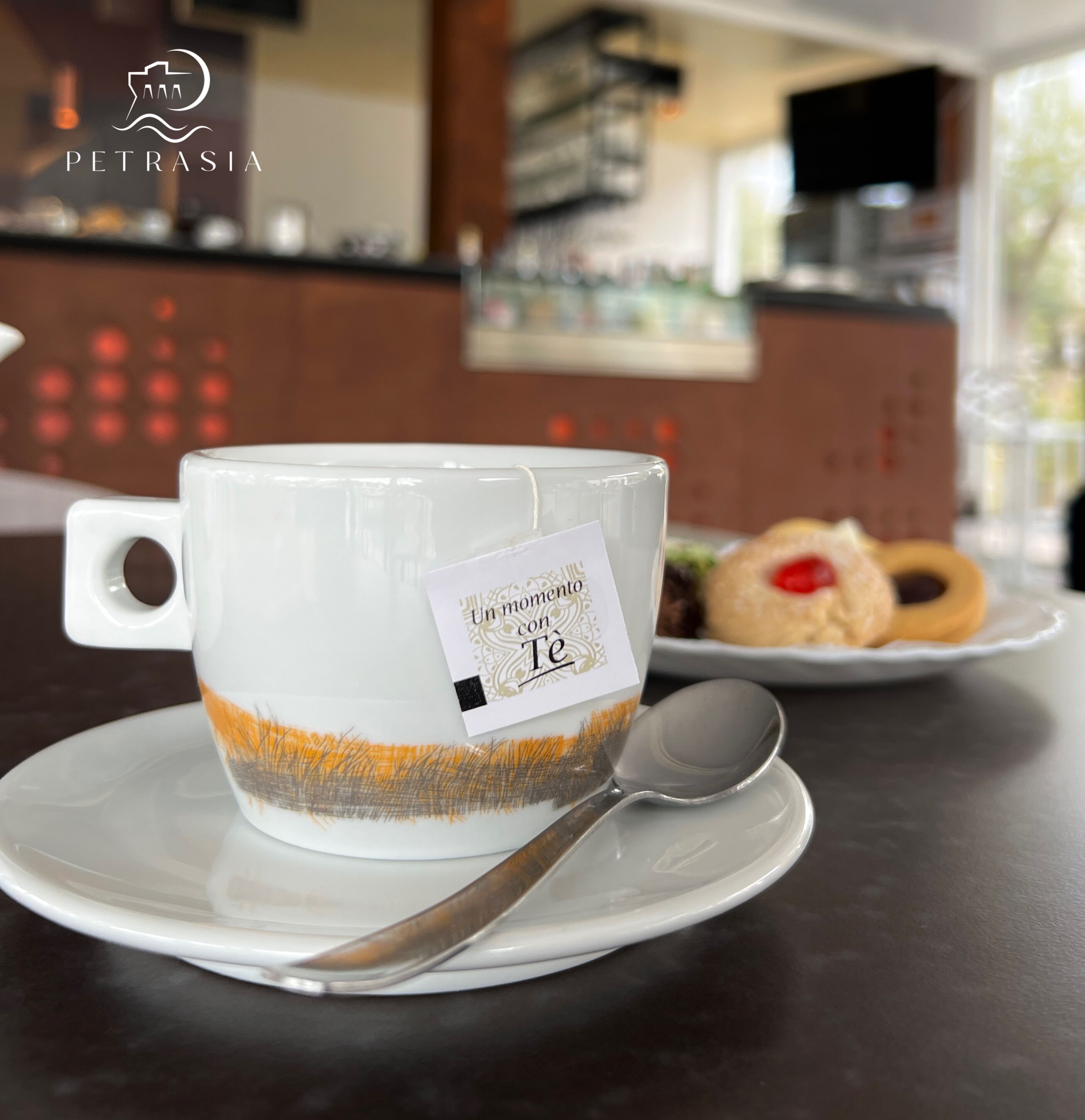 Une tasse blanche avec un sachet de thé posé sur une soucoupe, une cuillère et une assiette de pâtisseries, le tout dans un café.