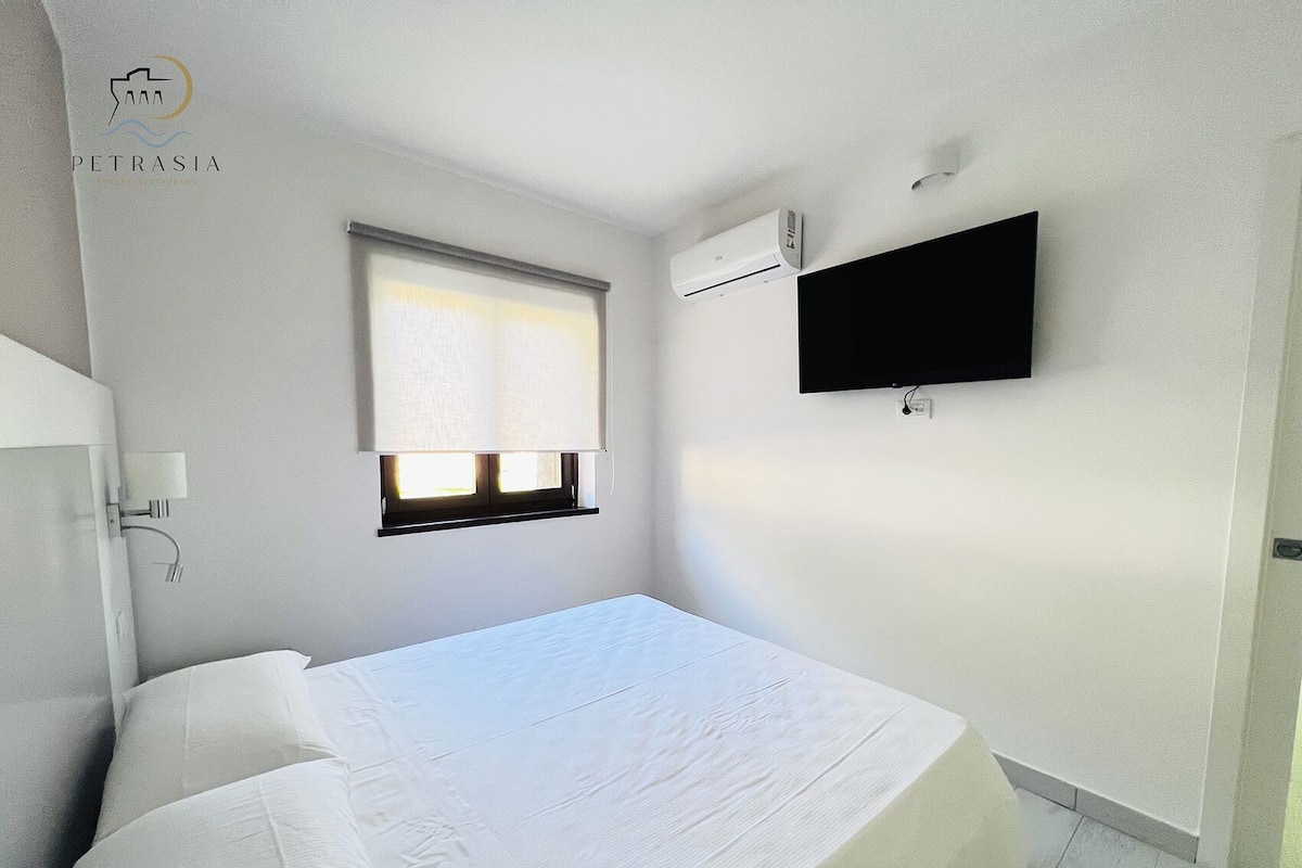 A bedroom with a bed, a window with blinds, a wall-mounted TV, and a ceiling-mounted air conditioner.