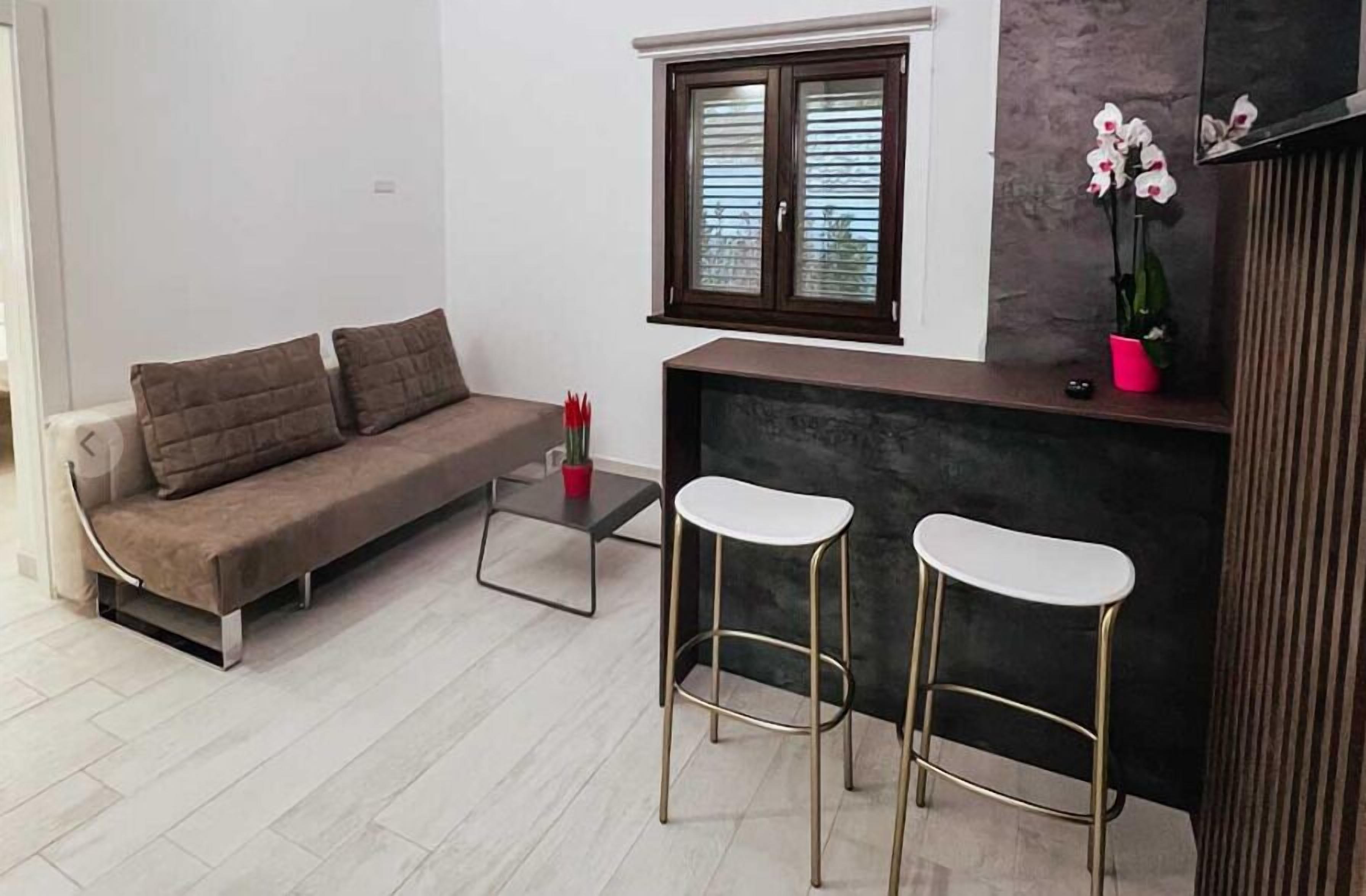 A modern living room with a sofa, two bar stools, a small table with a red vase, and a window with blinds.