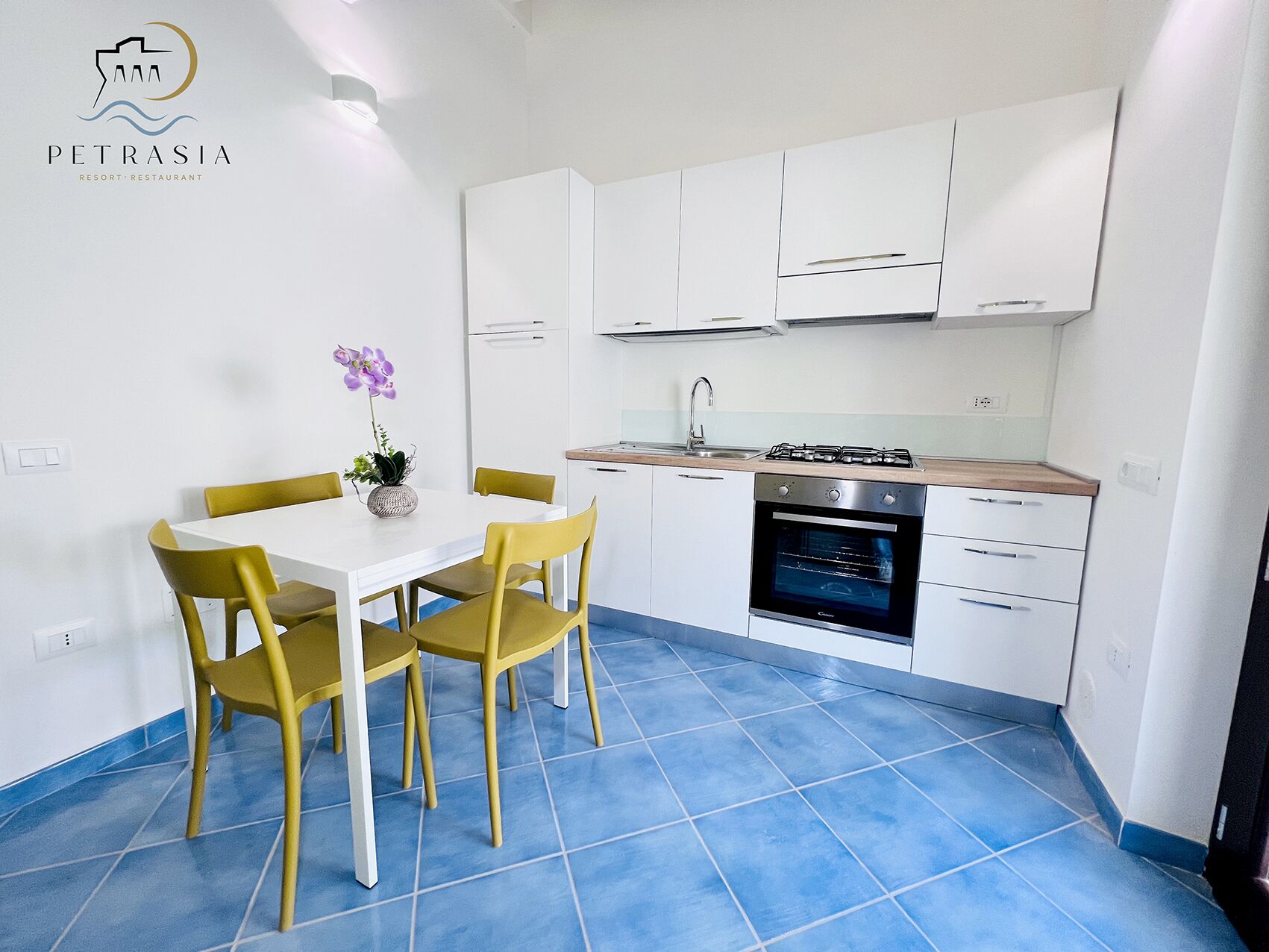 A modern kitchen with white cabinets, a gas stove, and a built-in oven. There is a dining table with yellow chairs and a vase with flowers.
