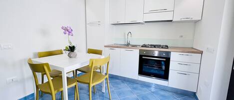 A modern kitchen with white cabinets, a gas stove, and a built-in oven. There is a dining table with yellow chairs and a vase with flowers.