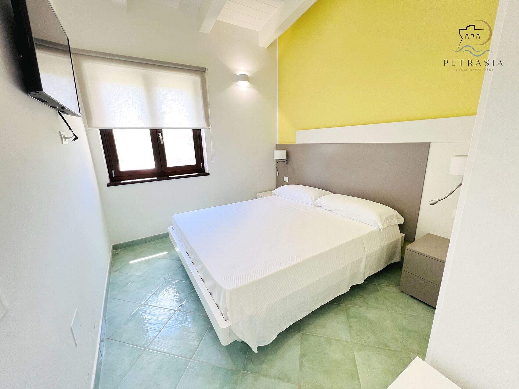 A bedroom with a bed, a nightstand, a wall-mounted lamp, a window with blinds, and a ceiling light.