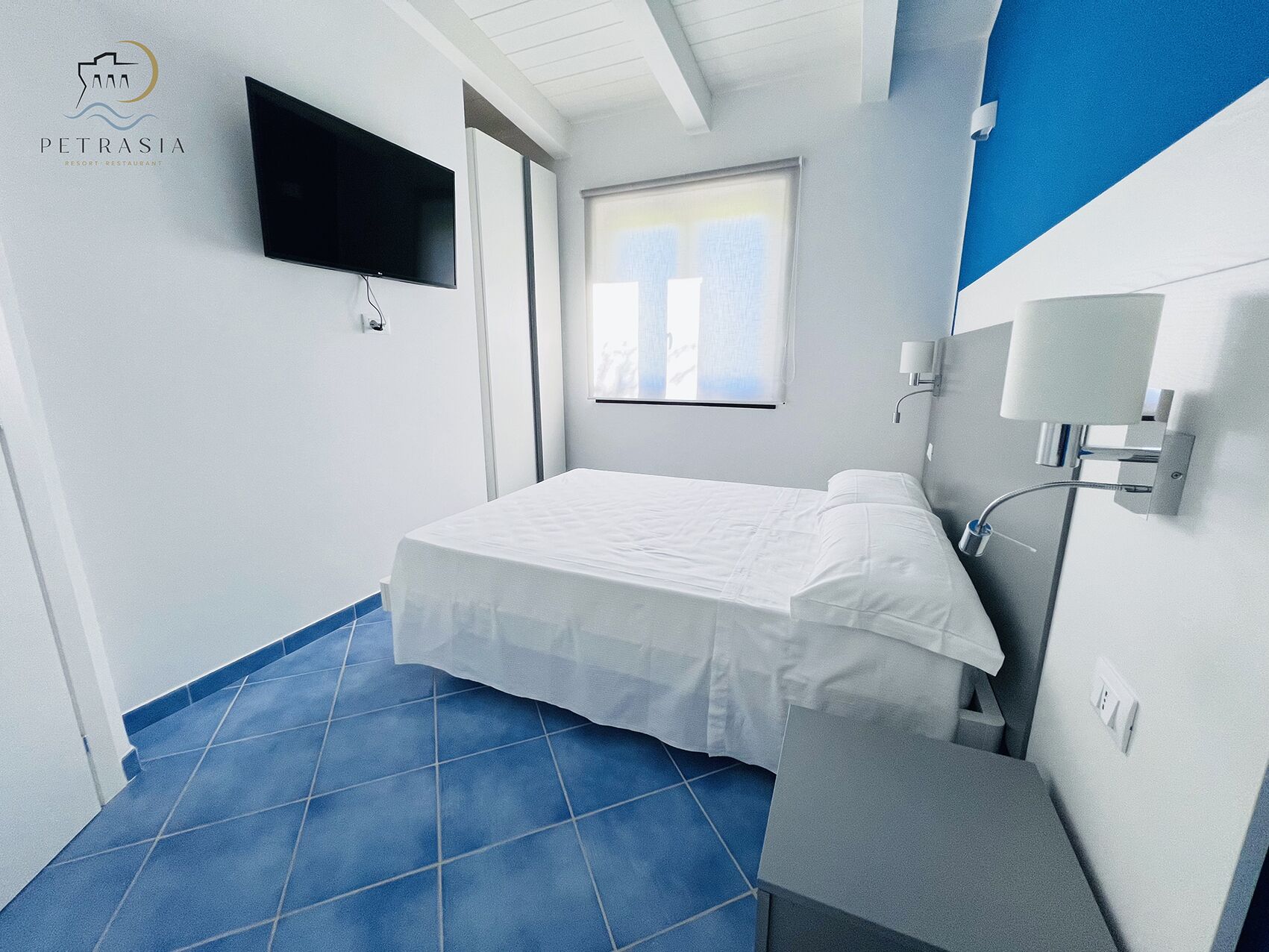 A hotel room with a bed, a TV mounted on the wall, a window with blinds, and a bathroom with a shower.