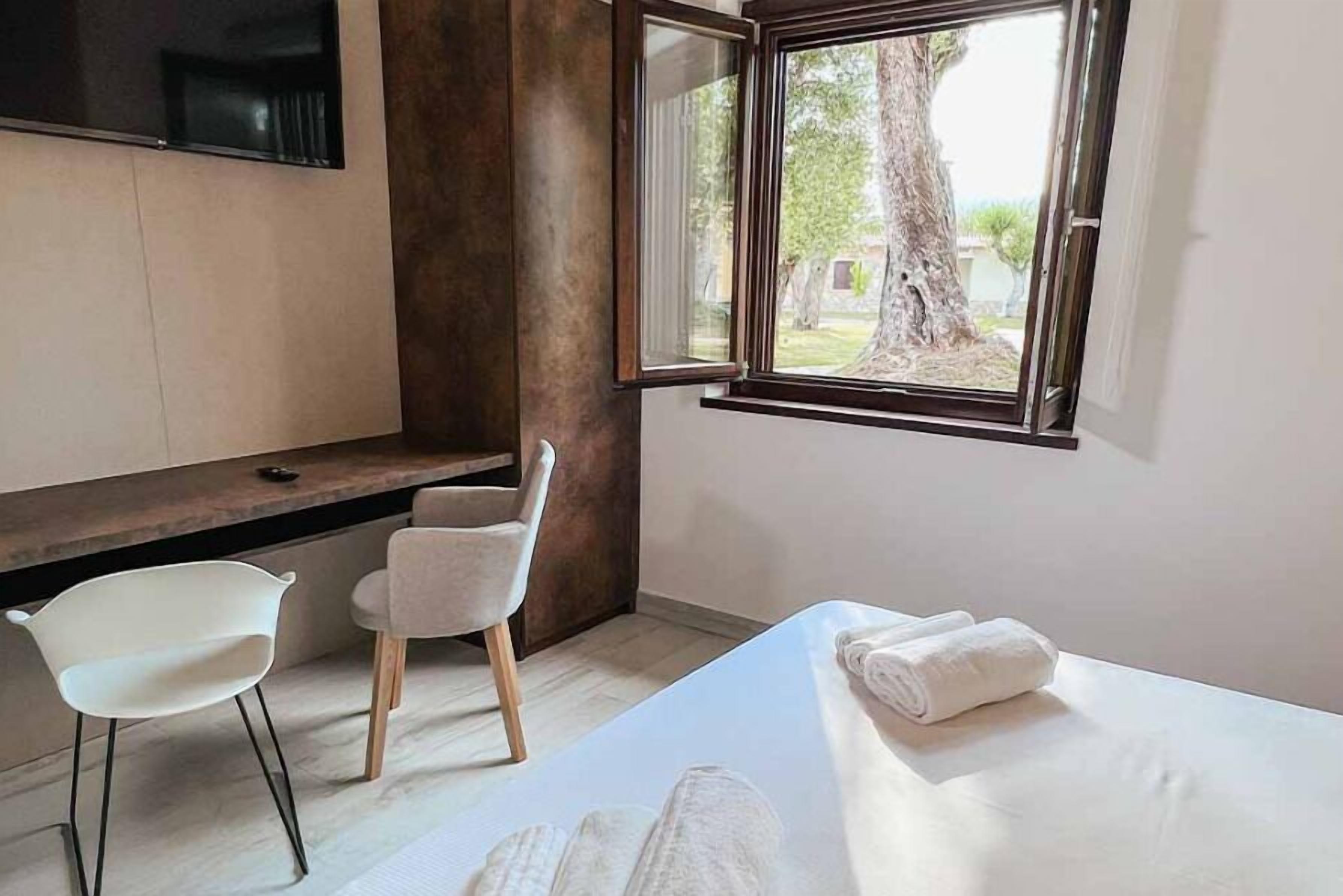 A modern hotel room with a bed, a desk, two chairs, and a large window with a view of trees.
