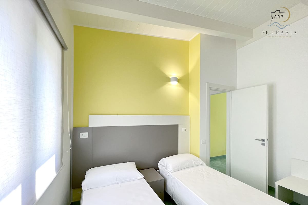 A hotel room with two single beds, a small bedside table, a nightstand, a wall-mounted light, and a door.