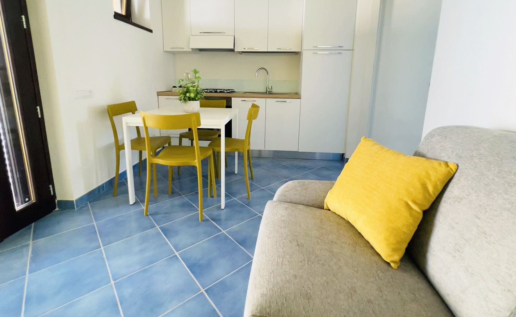 Un soggiorno con un divano color beige e un cuscino giallo, una zona pranzo con un tavolo bianco e sedie gialle, e una cucina con mobili bianchi e un fornello a gas.